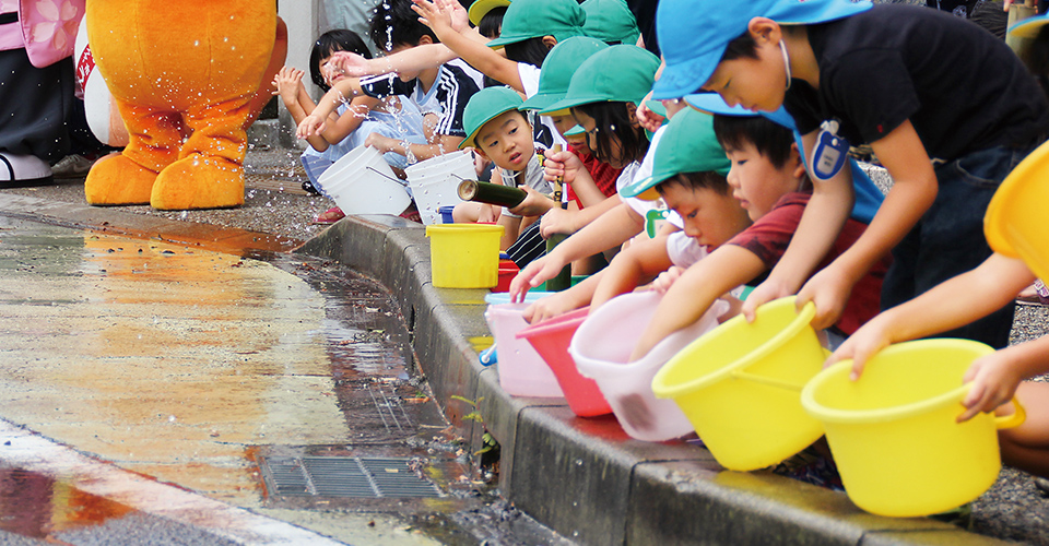 しゃがんだ子どもたちが一列になり、道路に水打ちしている様子（たじみクールアースデー）